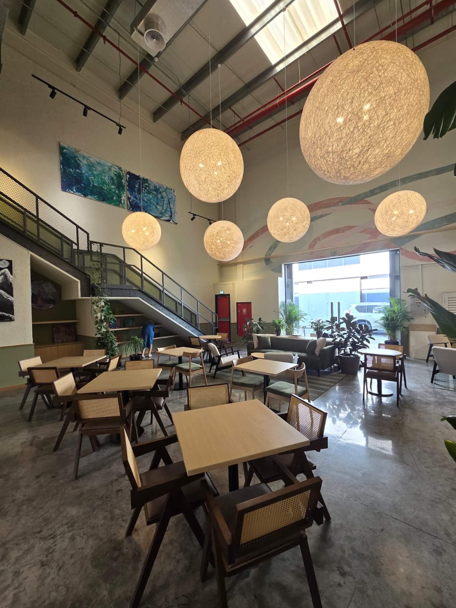 Modern cafe interior with hanging spherical lights, wooden tables, cane chairs, and high ceilings.