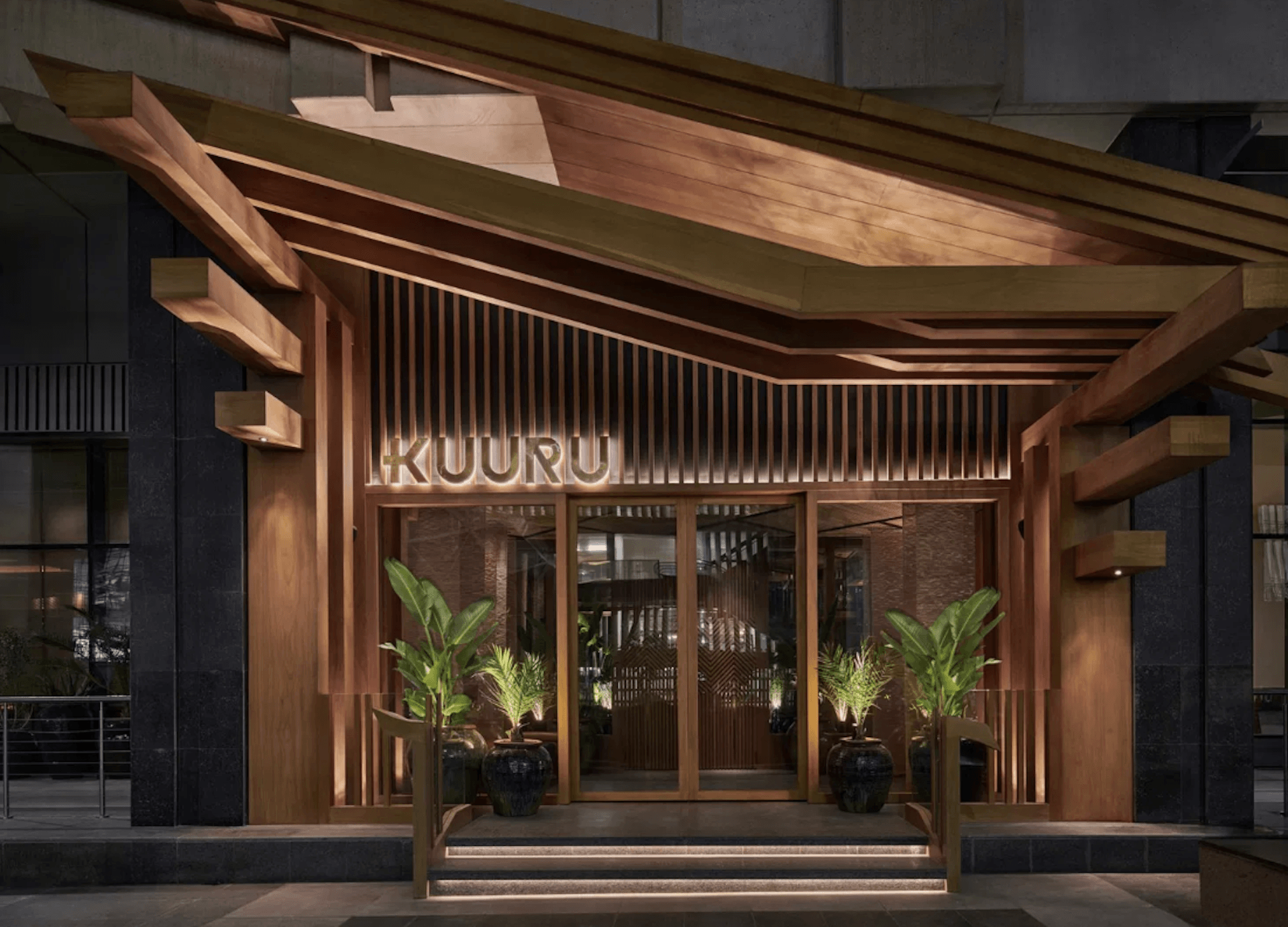 Modern restaurant storefront with illuminated "KUURU" sign, wooden pergola, and tropical plants