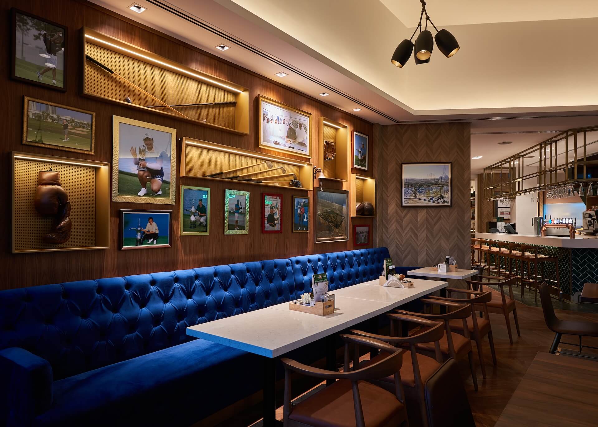 Modern sports bar with blue tufted seating, photo-lined walls, and illuminated wooden shelving.