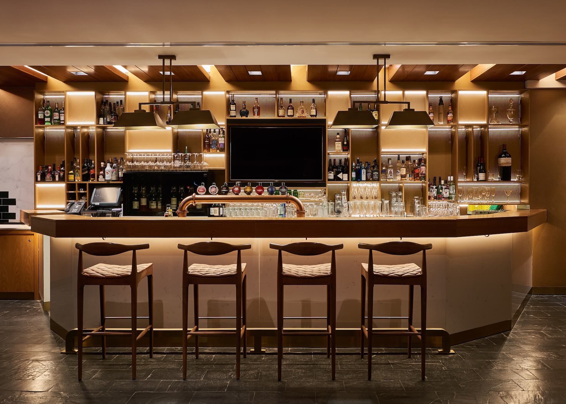 Modern luxury bar counter with four wooden stools, illuminated shelving displaying spirits bottles