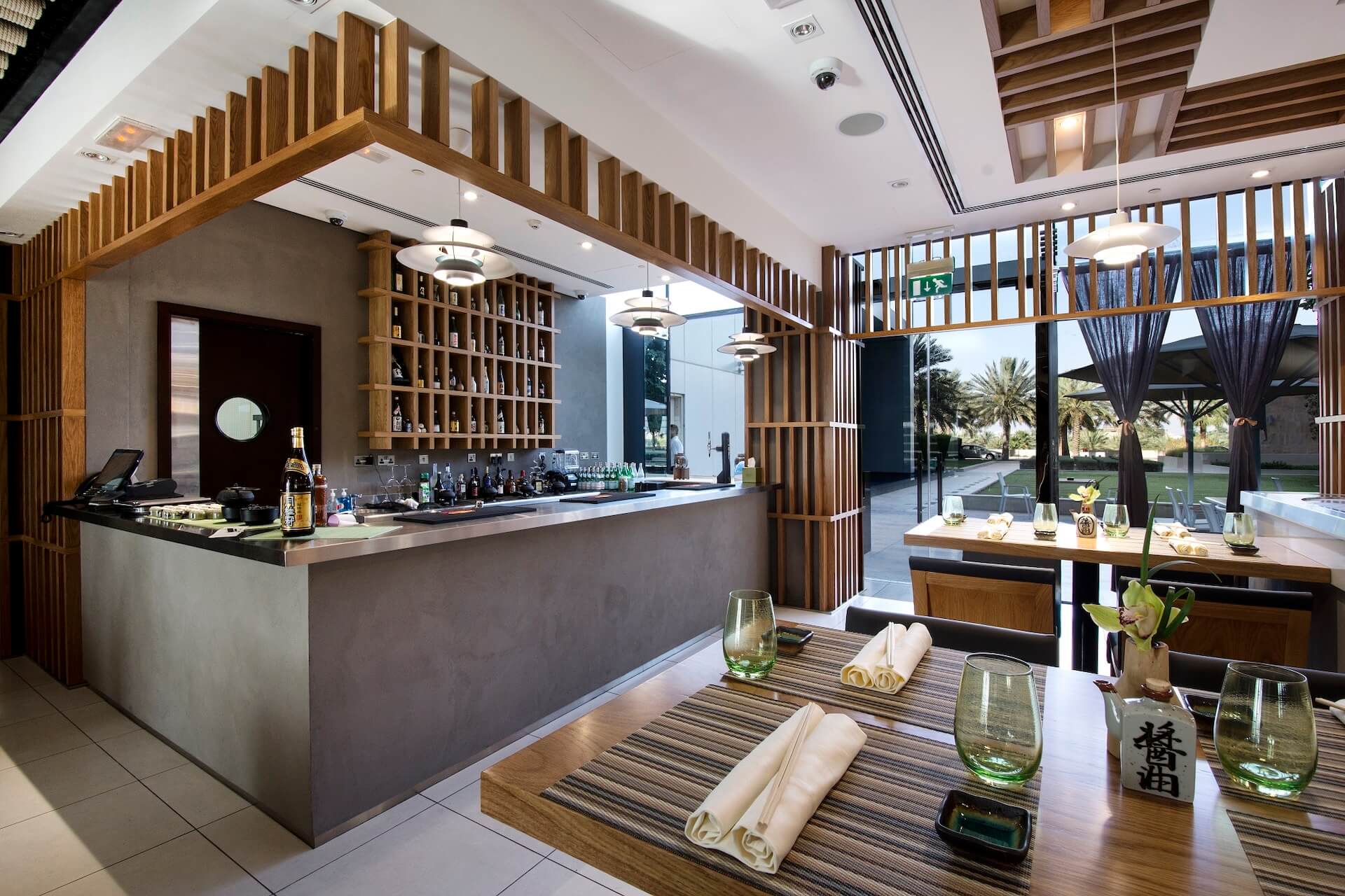 Modern spa reception area with wooden lattice screens, bar counter, and dining seating overlooking tropical landscape.