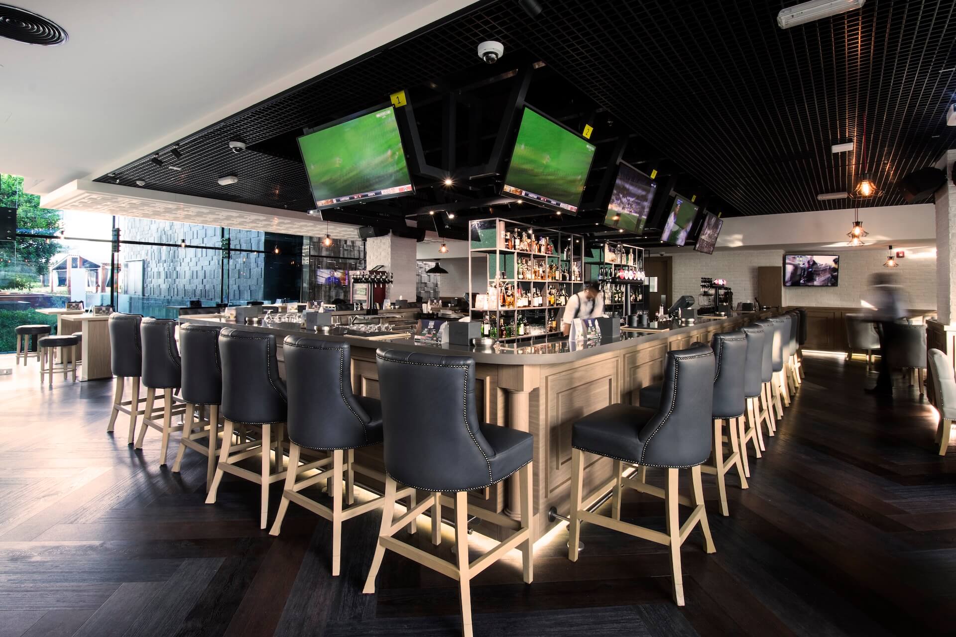 Modern bar interior with dark barstools, multiple TV screens, and well-stocked shelves