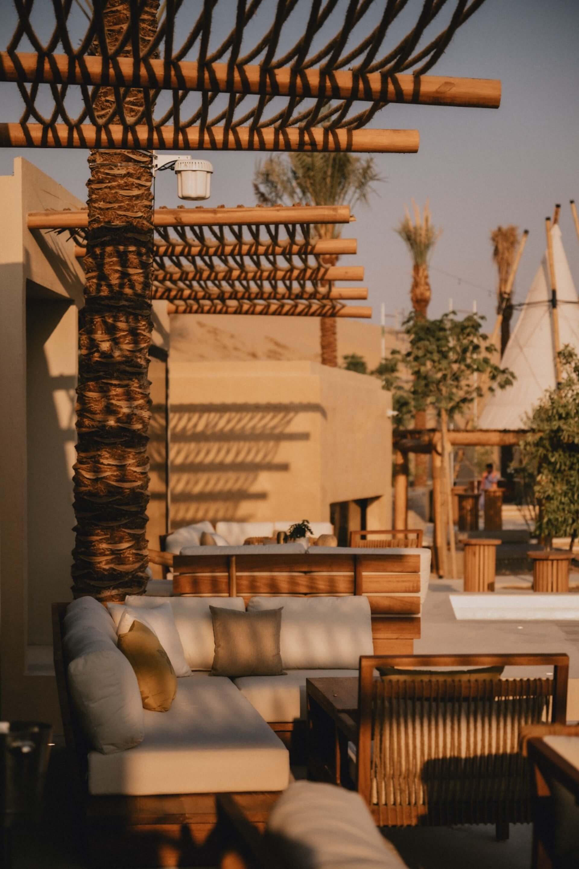 Luxury desert resort courtyard with palm tree, wooden pergola, white cushioned seating, and warm sunlight