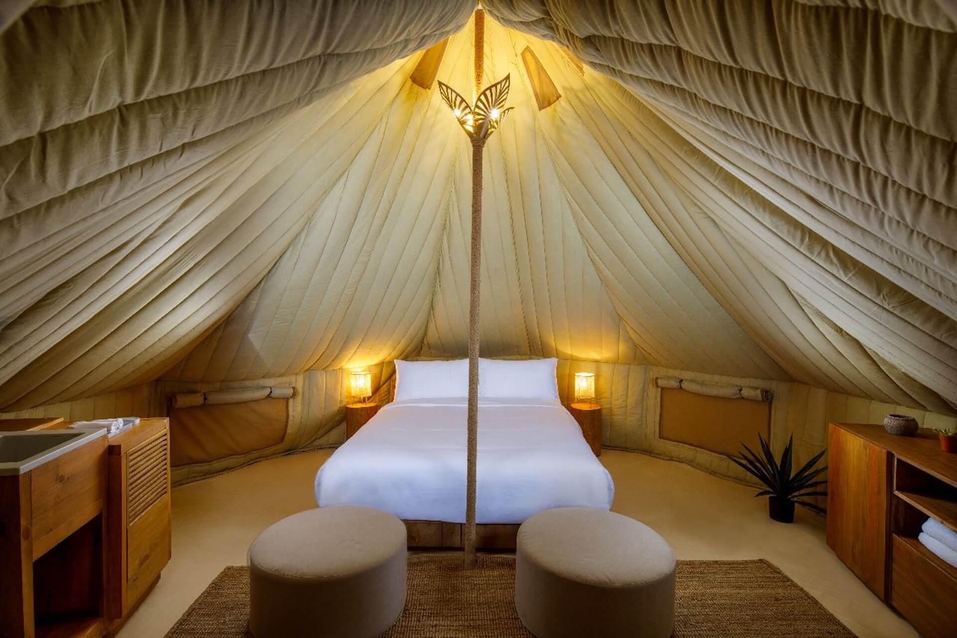 Luxury tent interior with white bed, wooden furniture, and golden lighting at apex