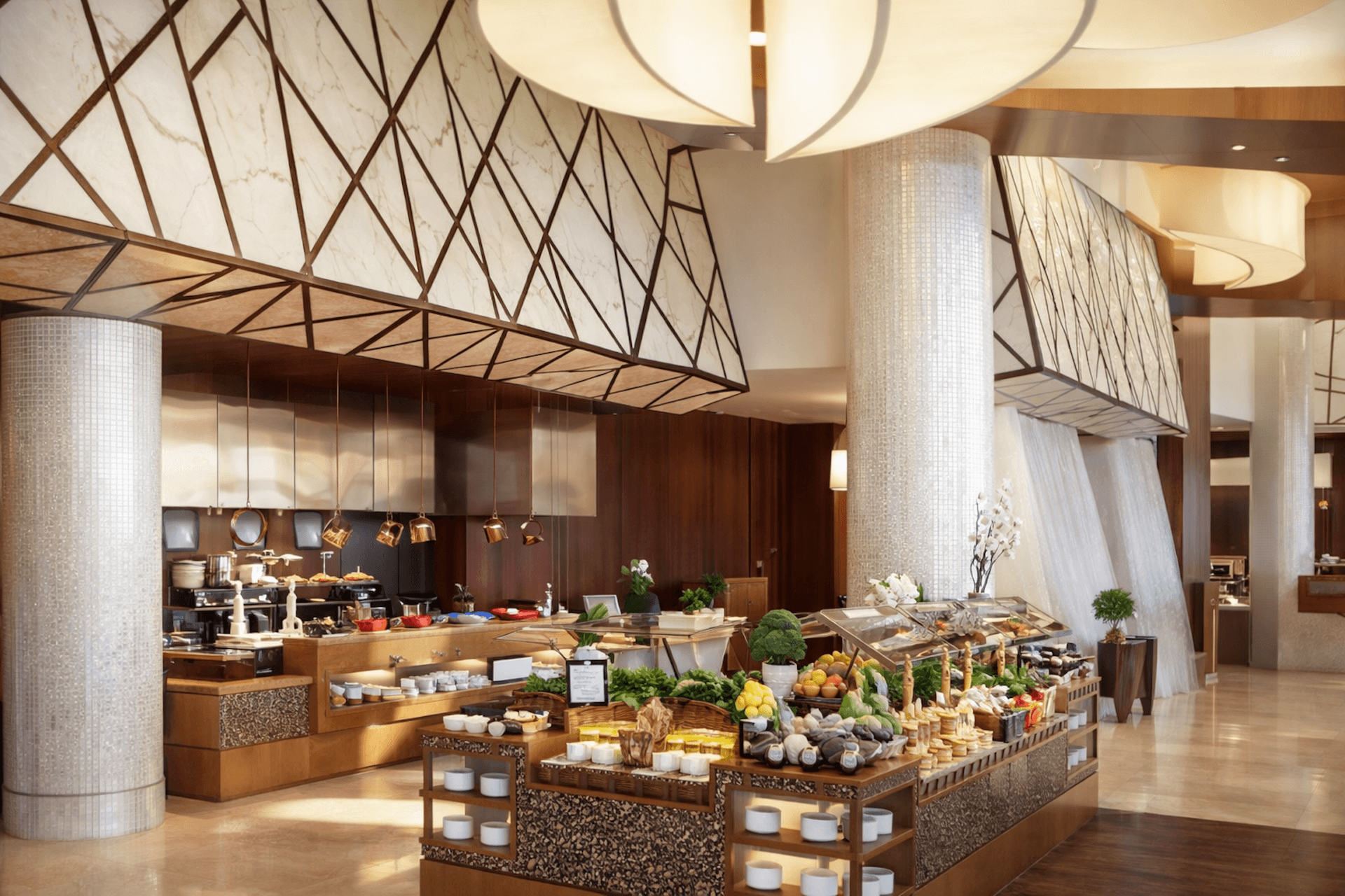 Modern hotel restaurant with open kitchen, fresh produce displays, and contemporary geometric ceiling design.