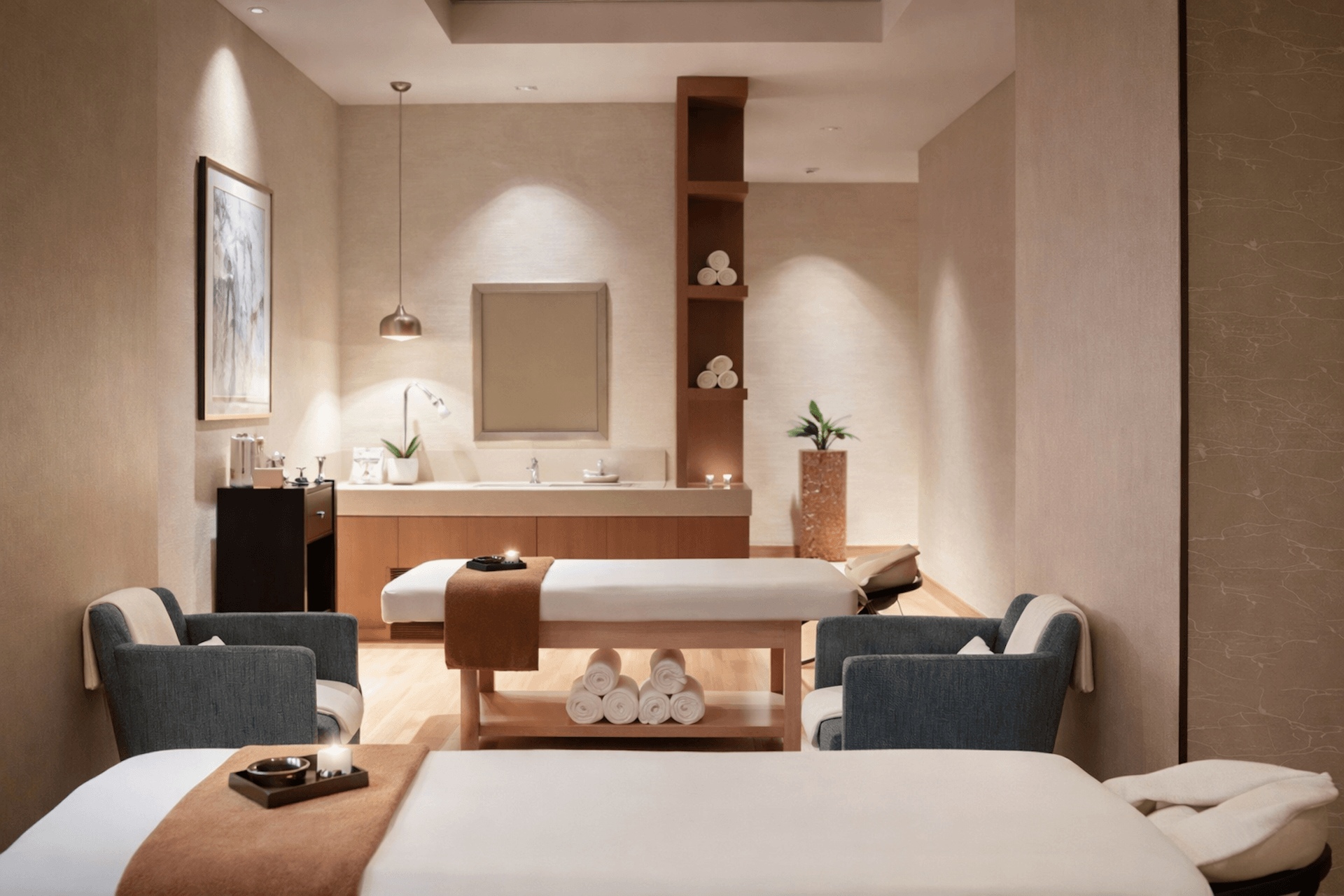 Modern spa treatment room with massage beds, sink vanity, shelving, plants, and warm lighting.