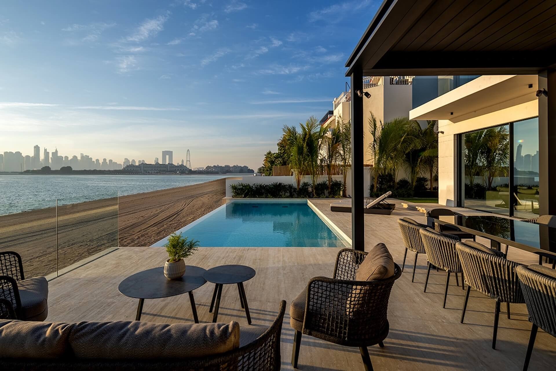 Modern waterfront mansion with infinity pool, patio seating, palm trees, and city skyline view.