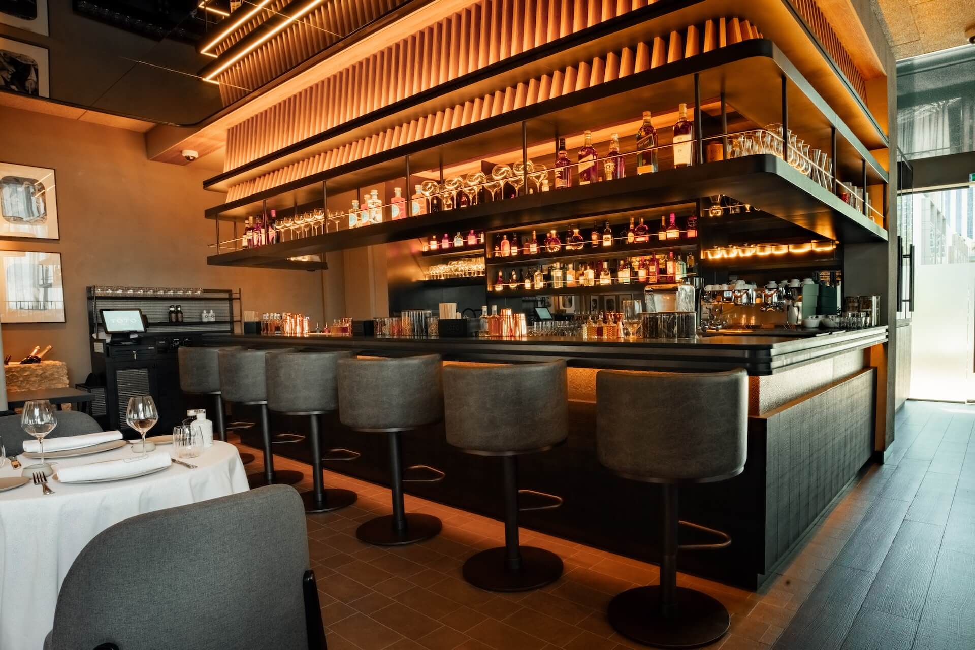 Modern upscale restaurant bar with warm lighting, bottle shelves, and dining area seating