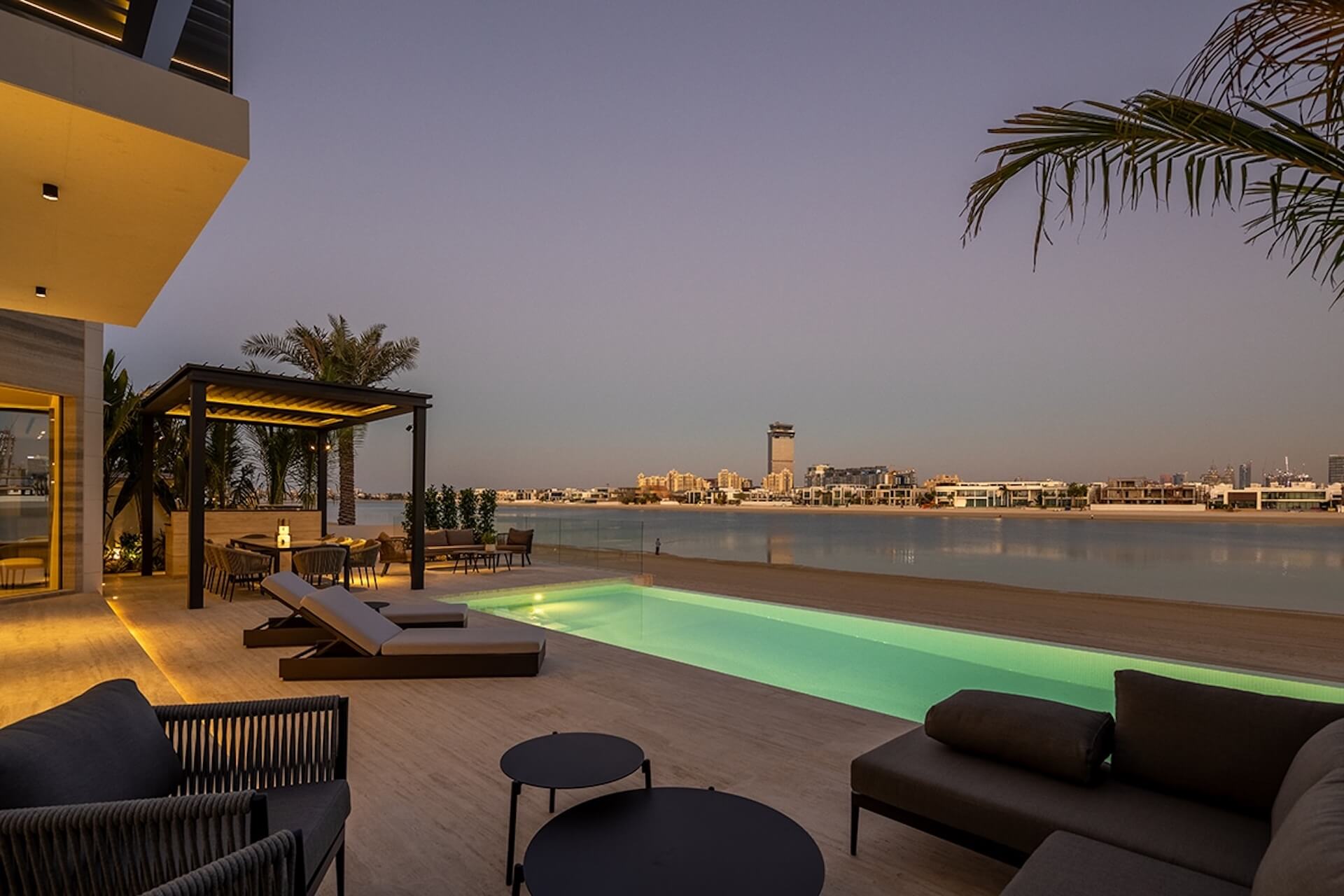 Luxury waterfront terrace at dusk with glowing pool, lounge furniture, and city skyline