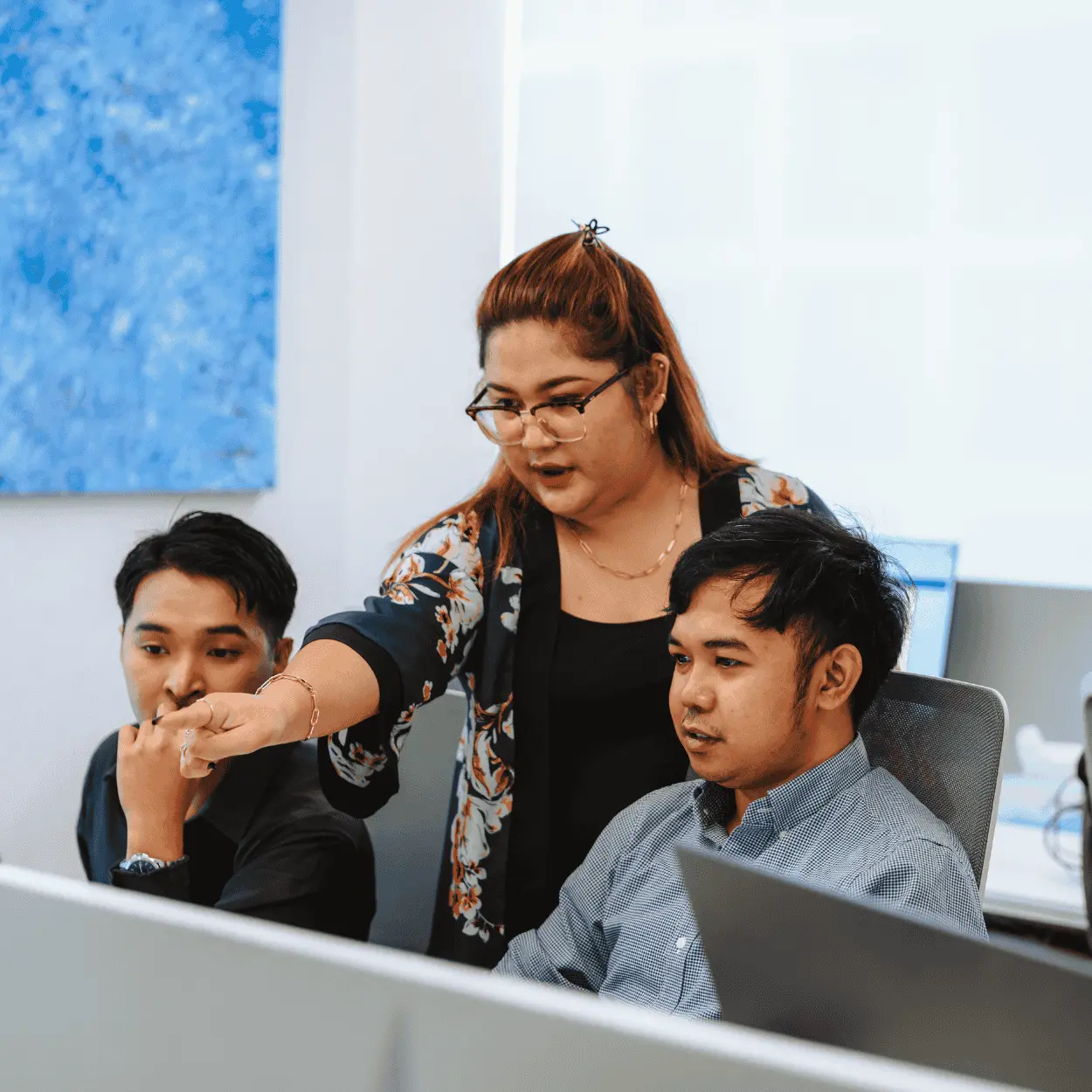 Woman in glasses explains work to two men at desk with laptop