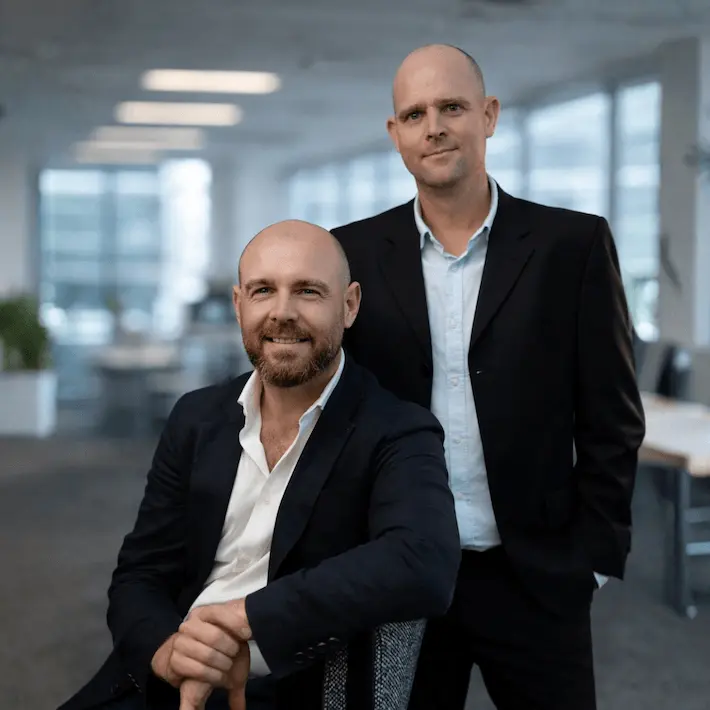 Two professional men in business suits posing in a modern office environment.