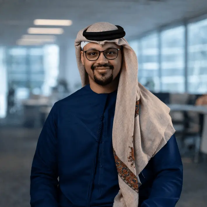Middle-aged man wearing traditional Arabic ghutra and agal, glasses, smiling in modern office setting.