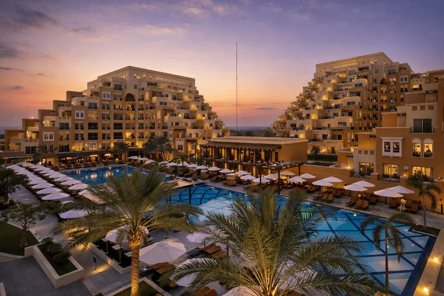 Luxury resort at dusk with illuminated buildings, pools, palm trees, and white lounge chairs.