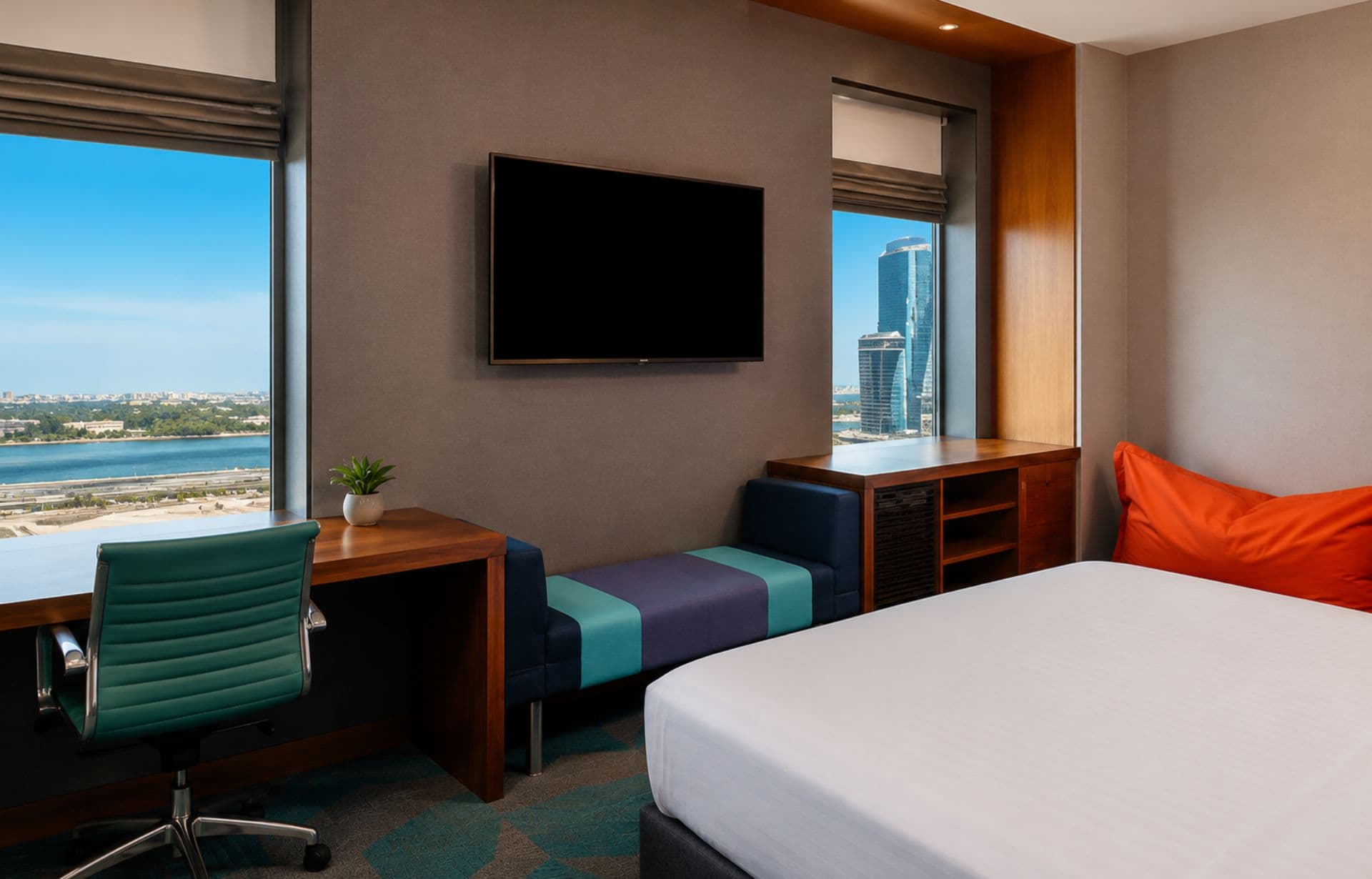 Modern hotel room with bed, desk, TV, and ocean city views from floor-to-ceiling windows