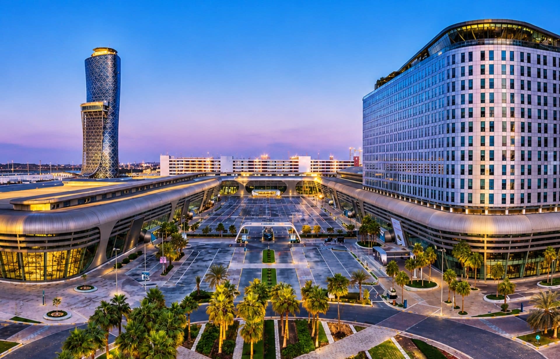 Modern urban plaza at twilight with illuminated towers, palm trees, and contemporary architecture