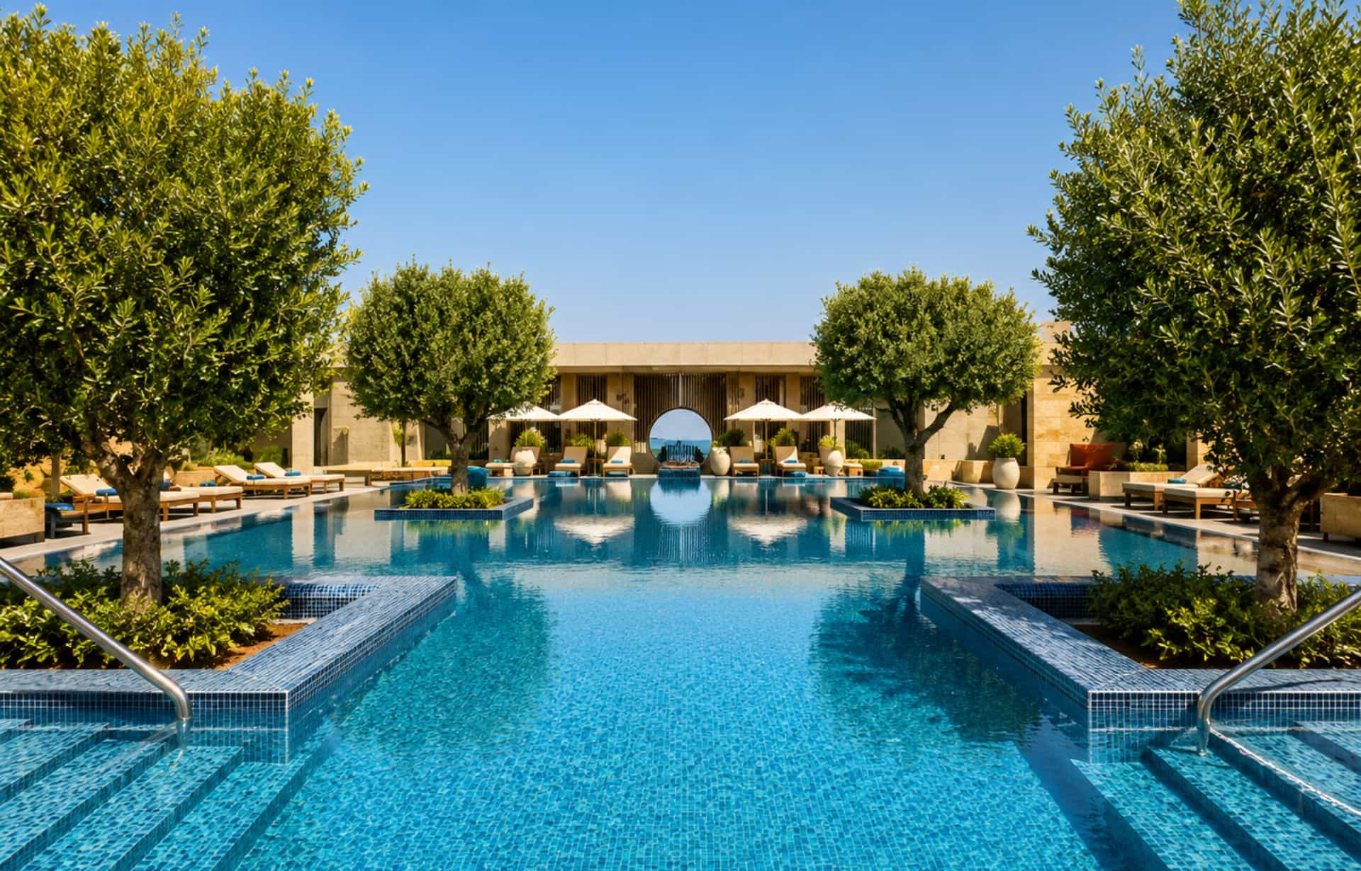 Luxurious resort courtyard with turquoise pool, loungers, umbrellas, olive trees, and tan stone building.