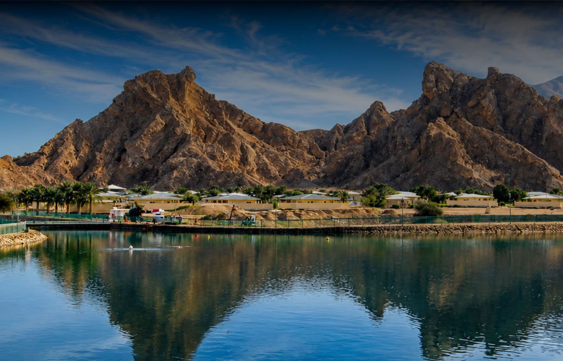 Dramatic rocky mountains reflect in calm river with palm trees and yellow buildings below under blue sky