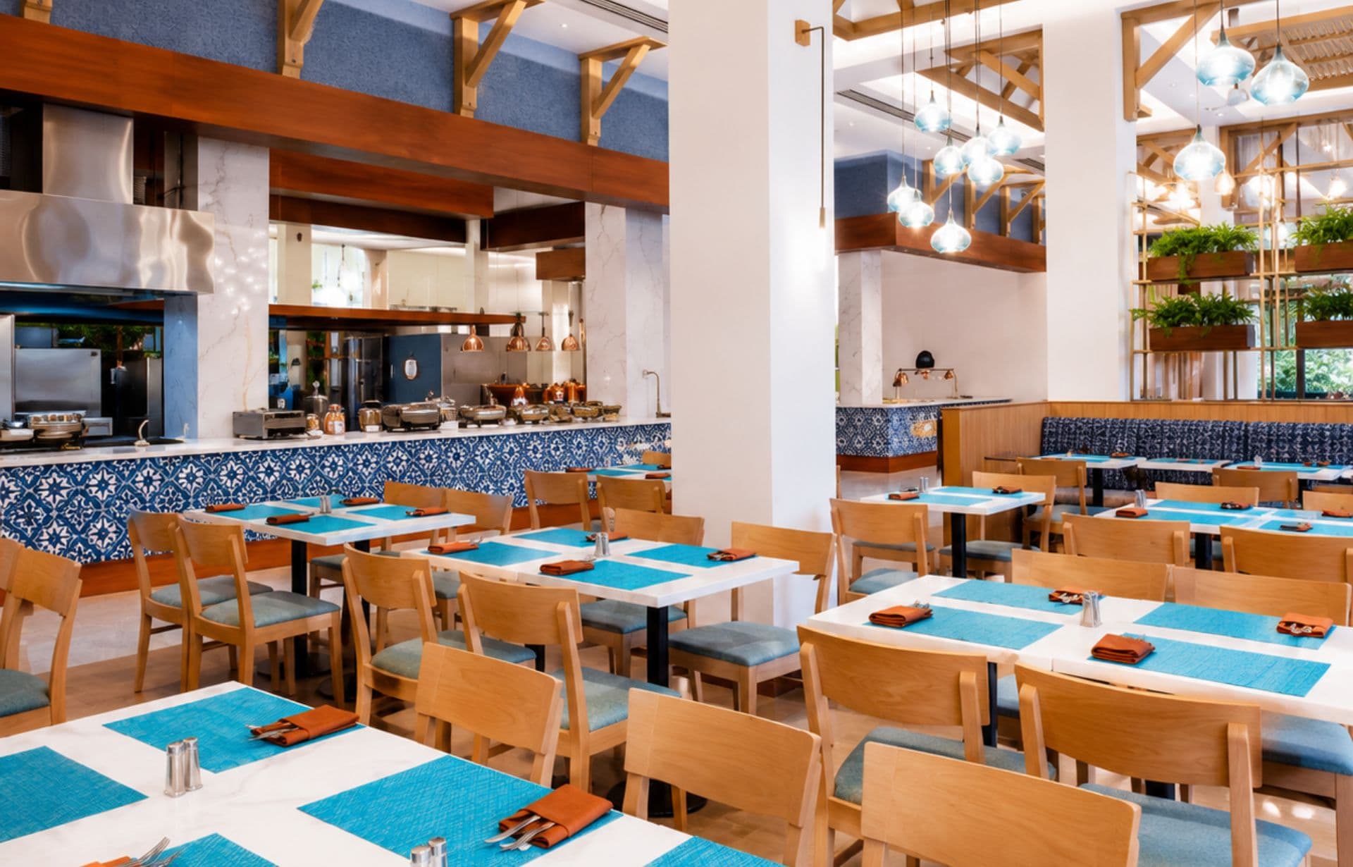 Modern restaurant interior with wooden tables, blue place settings, pendant lights, and open kitchen.