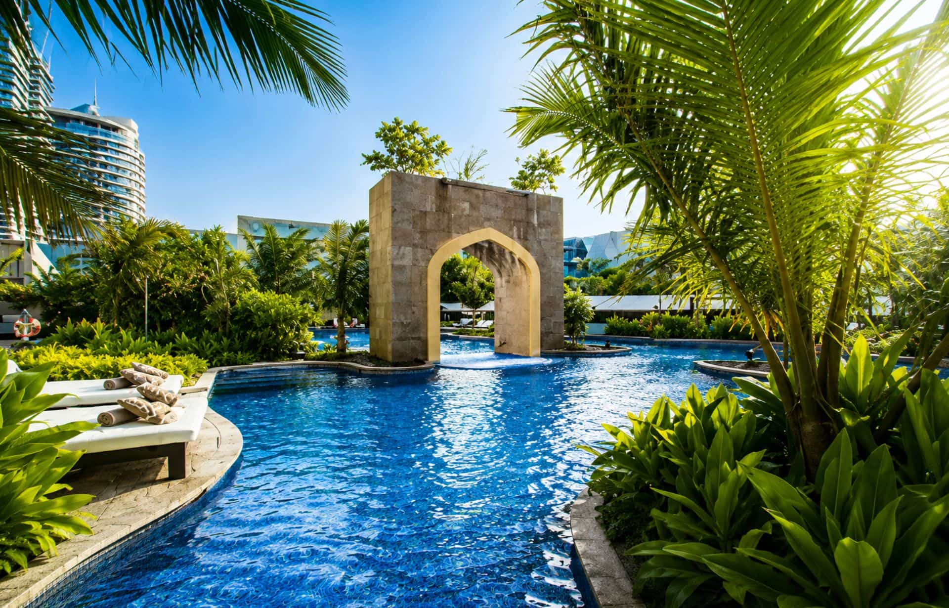 Luxury resort pool with tropical plants, archway feature, and modern high-rise buildings under blue sky