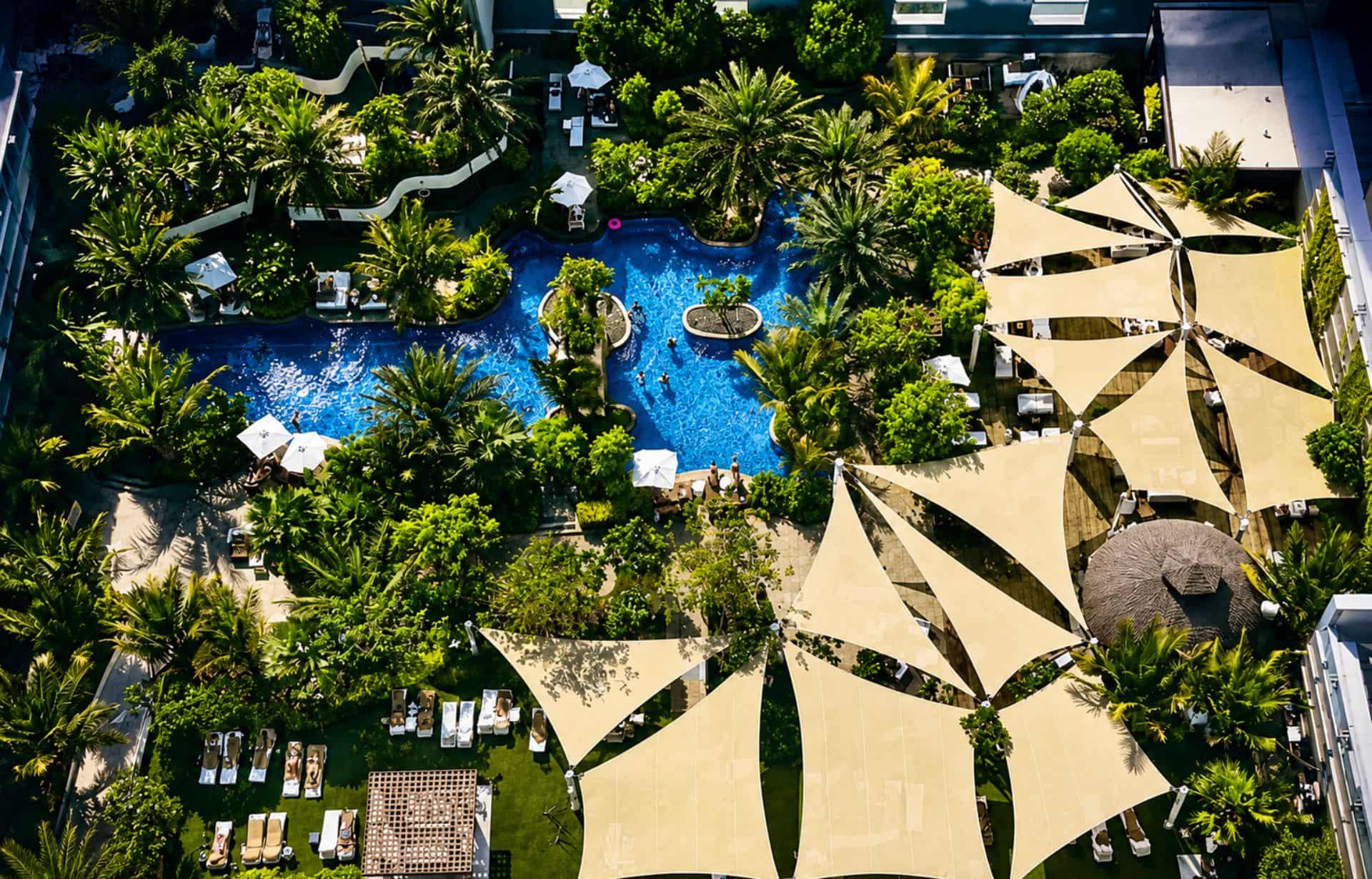 Aerial view of luxury resort with blue swimming pool, tan shade sails, and tropical landscaping.