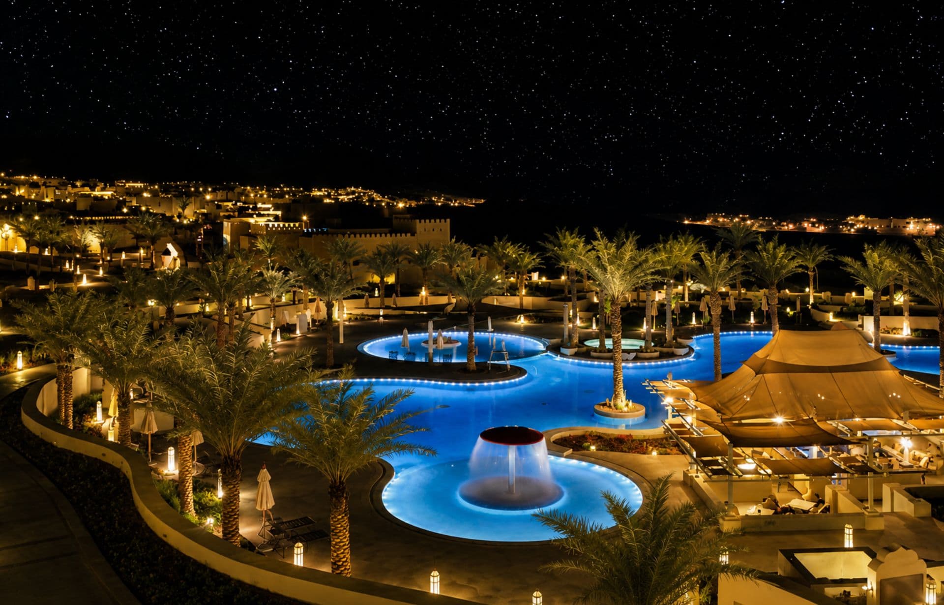 Luxurious resort with glowing blue pools, palm trees, and ancient fortress under starry night sky.