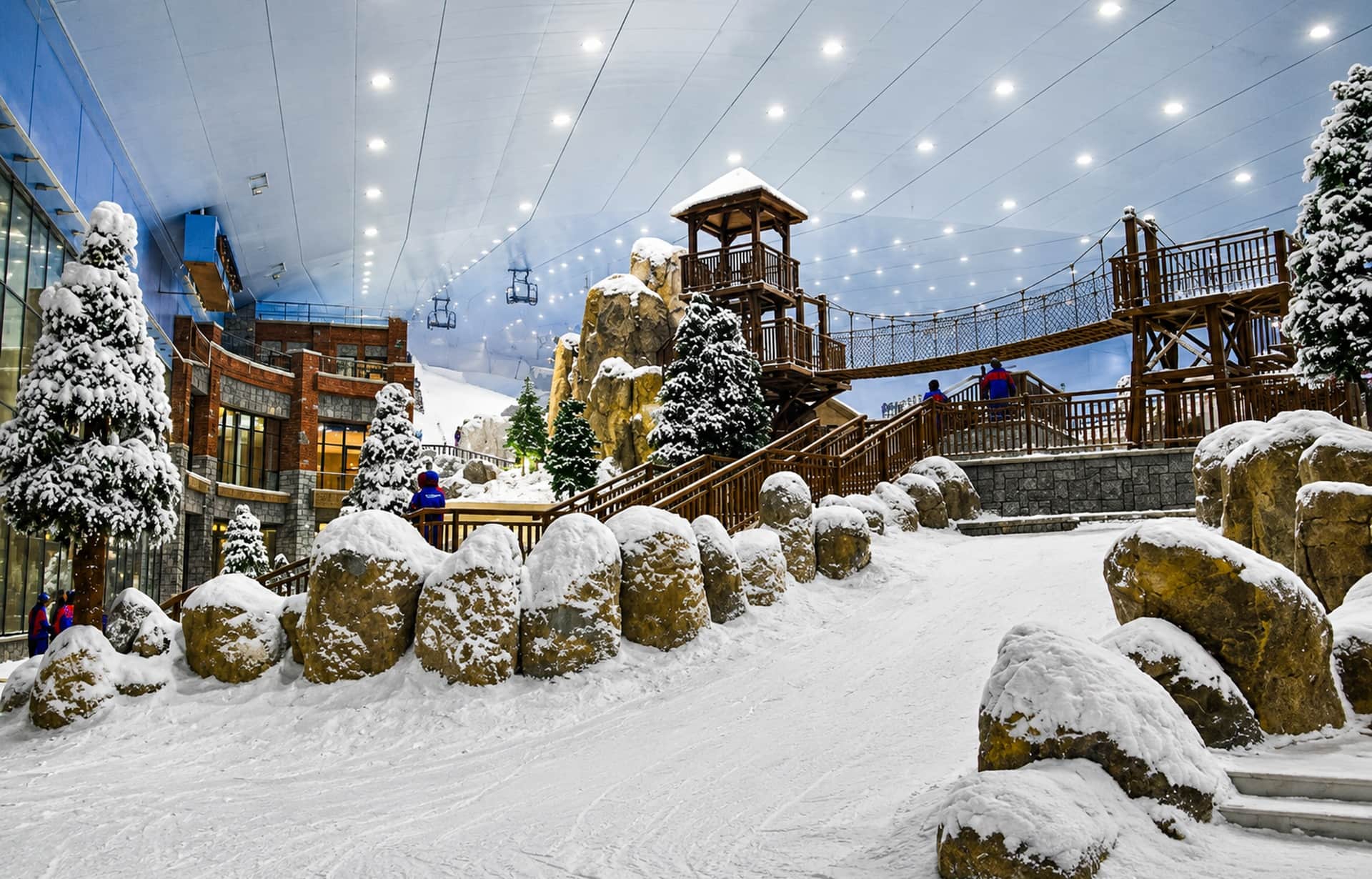 Indoor ski resort with snow-covered trees, wooden structures, and artificial snowfall under bright ceiling lights.