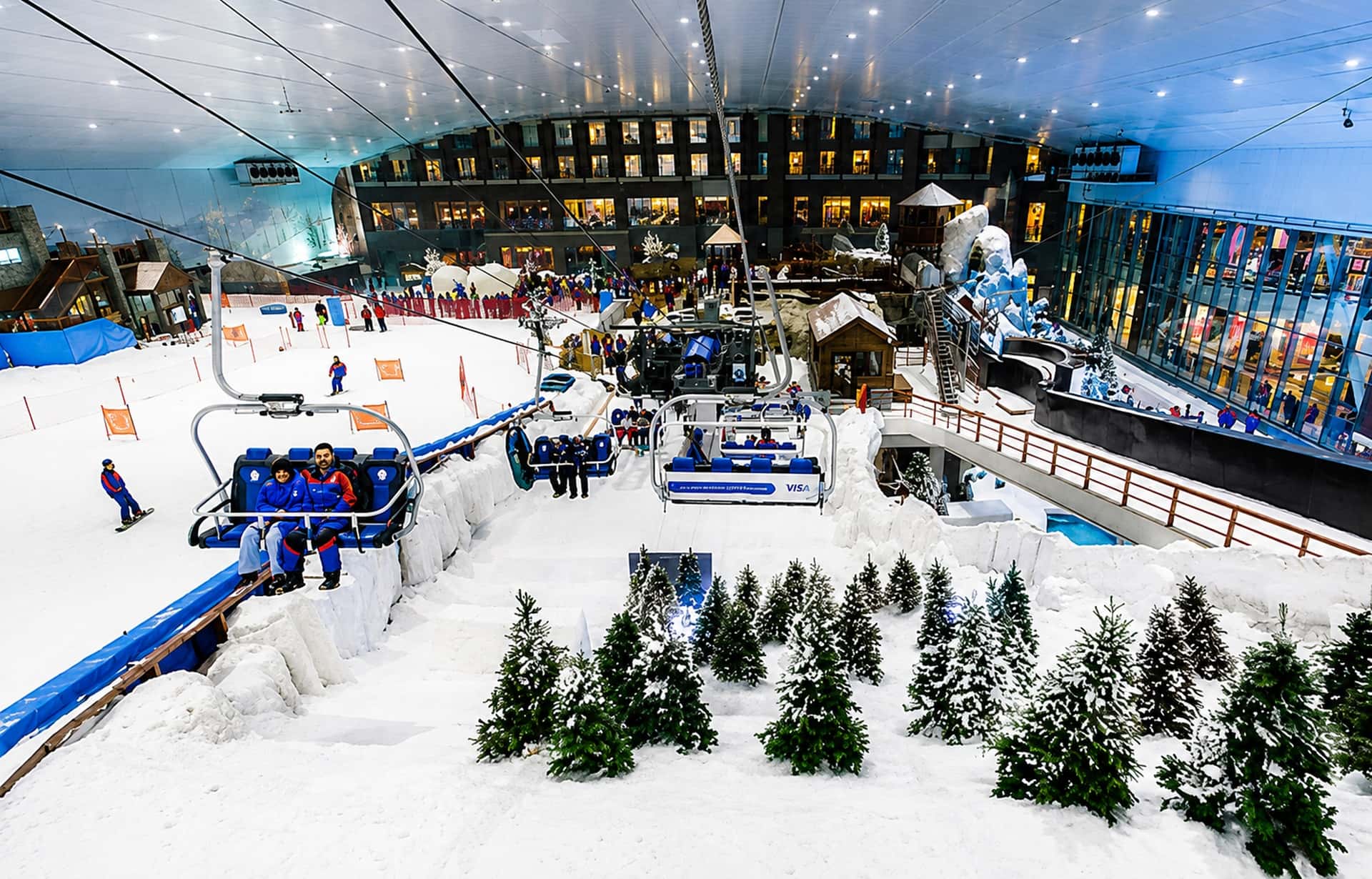 Indoor ski resort with snowy slopes, rides, visitors, and alpine trees under artificial lighting.