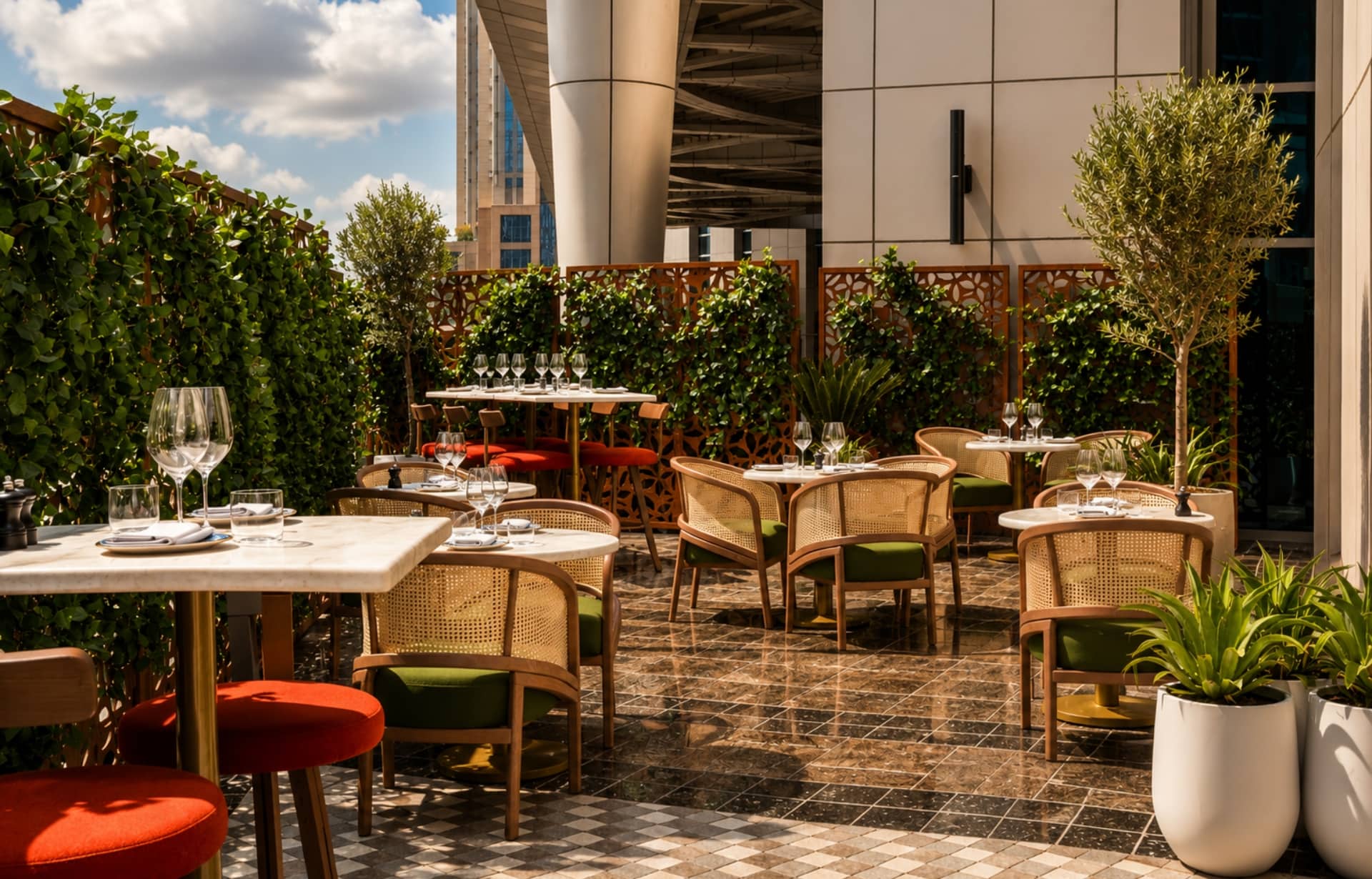 Modern rooftop restaurant terrace with dining tables, rattan chairs, lush green plants, and urban skyline view.