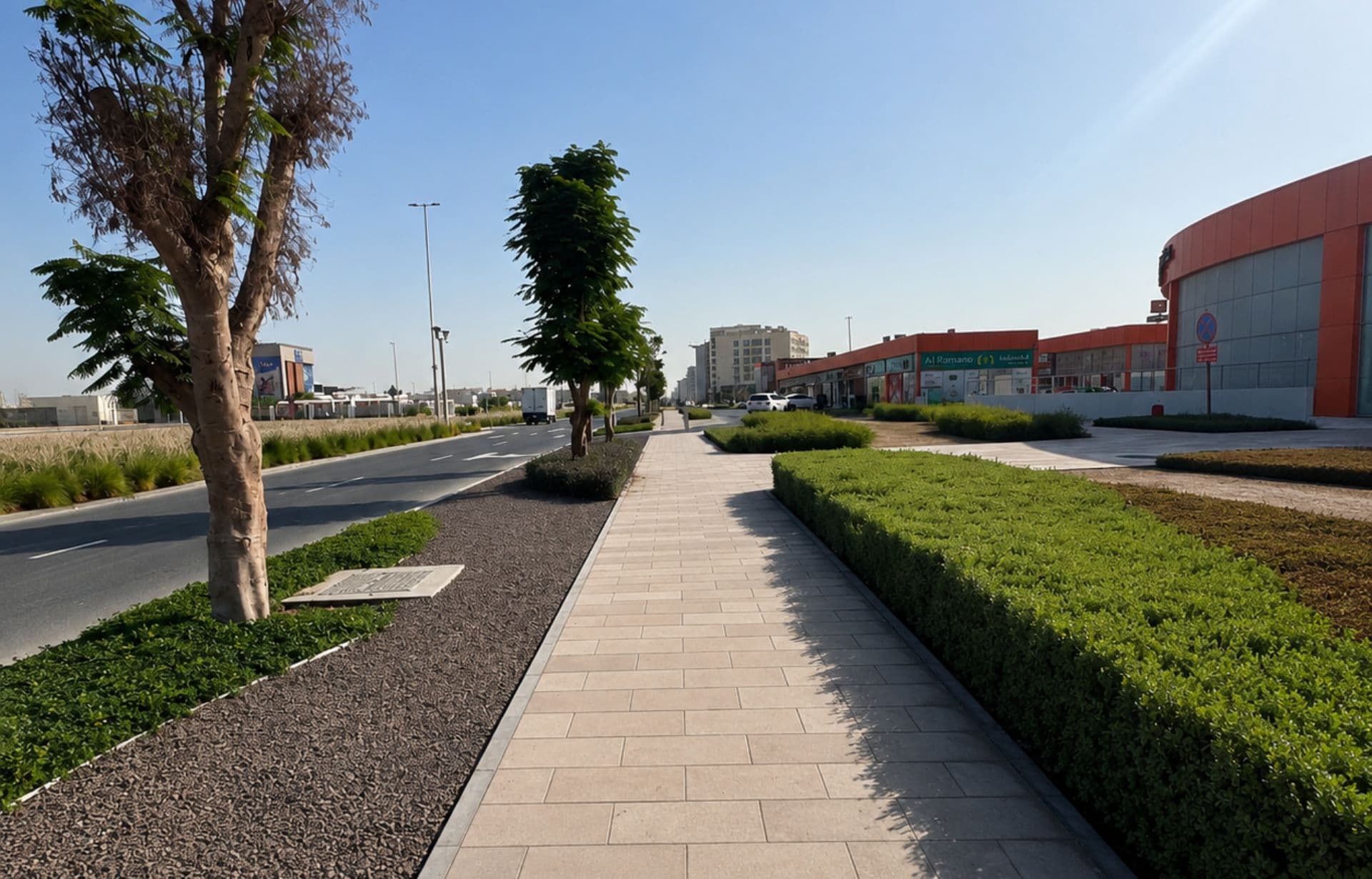 Modern suburban street with pedestrian pathway, trees, landscaping, and commercial buildings under clear sky