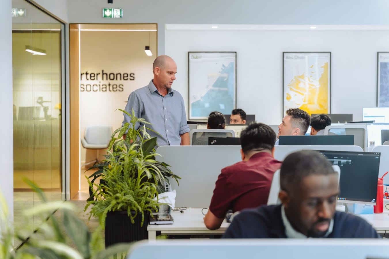 Man in blue shirt presenting to seated colleagues in modern office with computers and green plants.