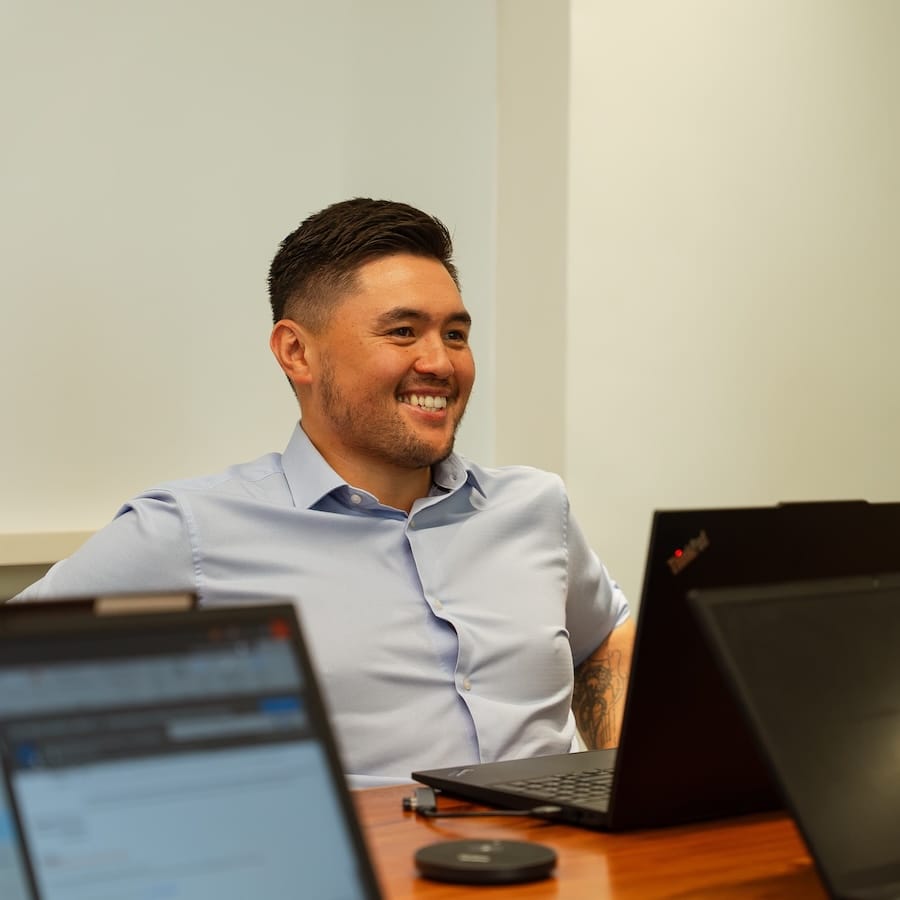Smiling man in light blue shirt at desk with multiple computers and laptops