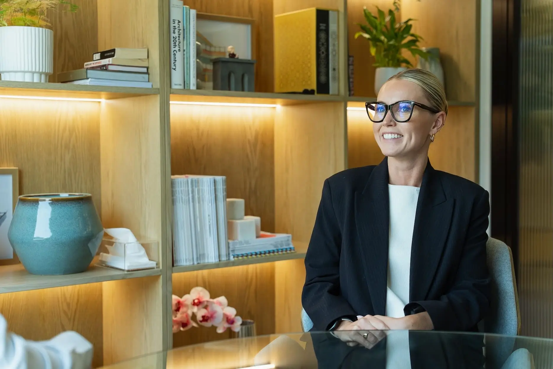 Professional woman in glasses and blazer sits at desk with wooden shelving unit backdrop.