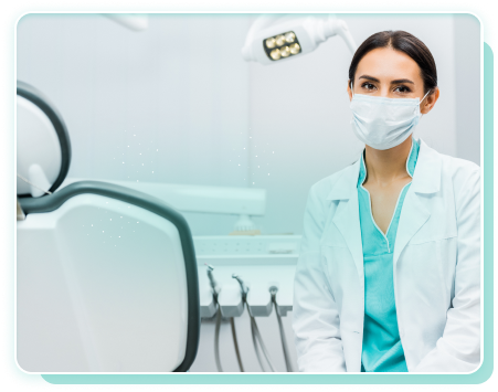 Female dentist wearing a surgical mask and white coat sitting in a dental clinic with dental chair and equipment in the background.