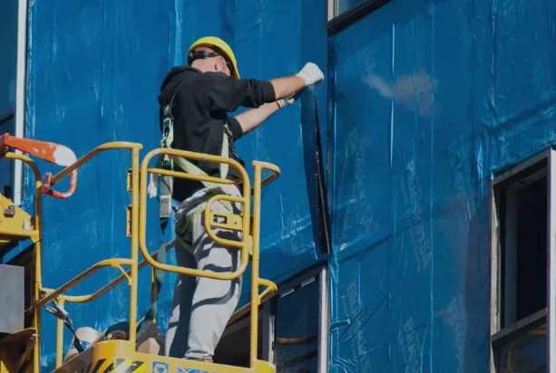 Construction worker in a yellow helmet and safety harness working on blue exterior insulation on a building from a yellow lift platform.