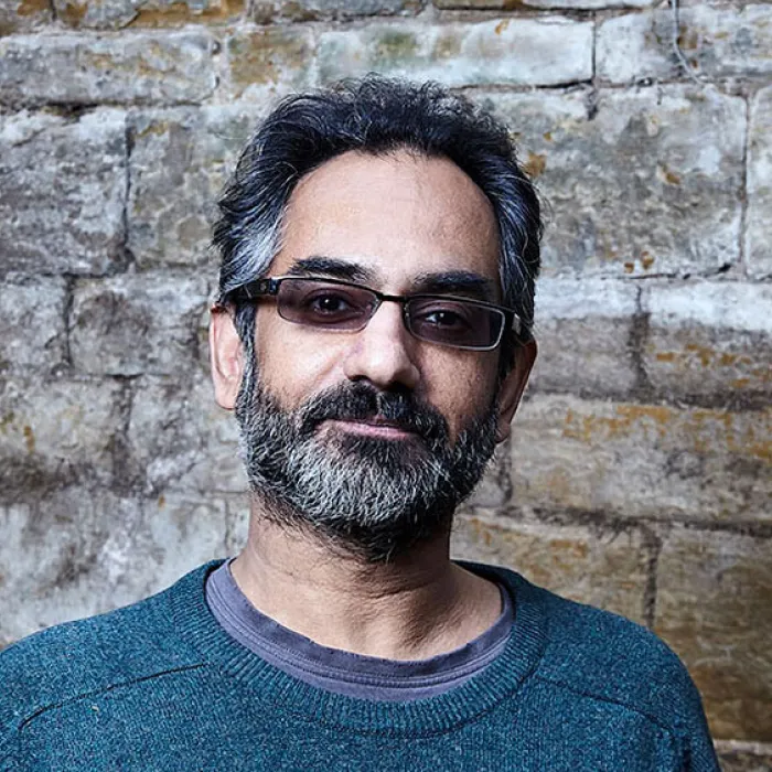 Middle-aged man with salt-and-pepper hair and beard wearing dark glasses and a teal sweater, standing in front of a stone wall.
