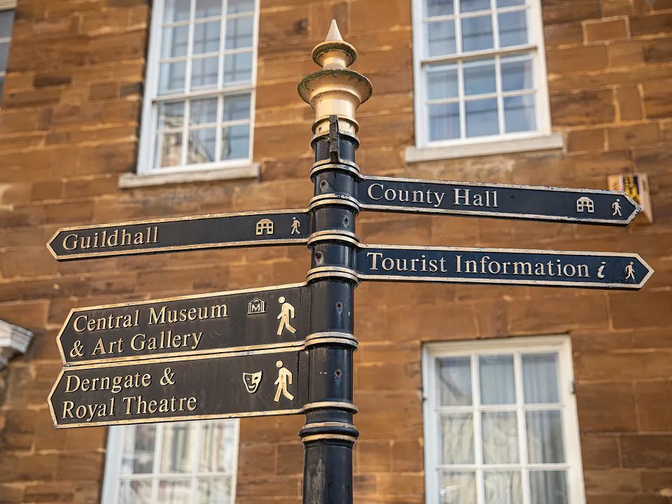 Black metal signpost with gold text pointing to Guildhall, County Hall, Tourist Information, Central Museum & Art Gallery, and Derngate & Royal Theatre against a brick building with white-framed windows.