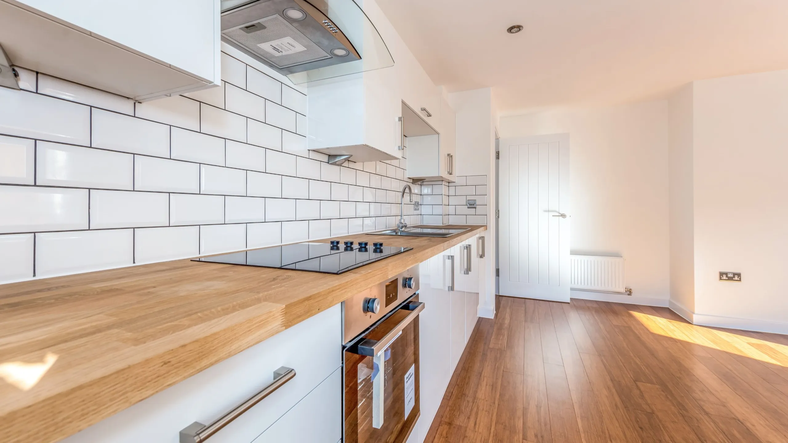 Modern kitchen with wooden countertop, white cabinets, built-in oven, electric stovetop, white subway tile backsplash, and wooden floor.