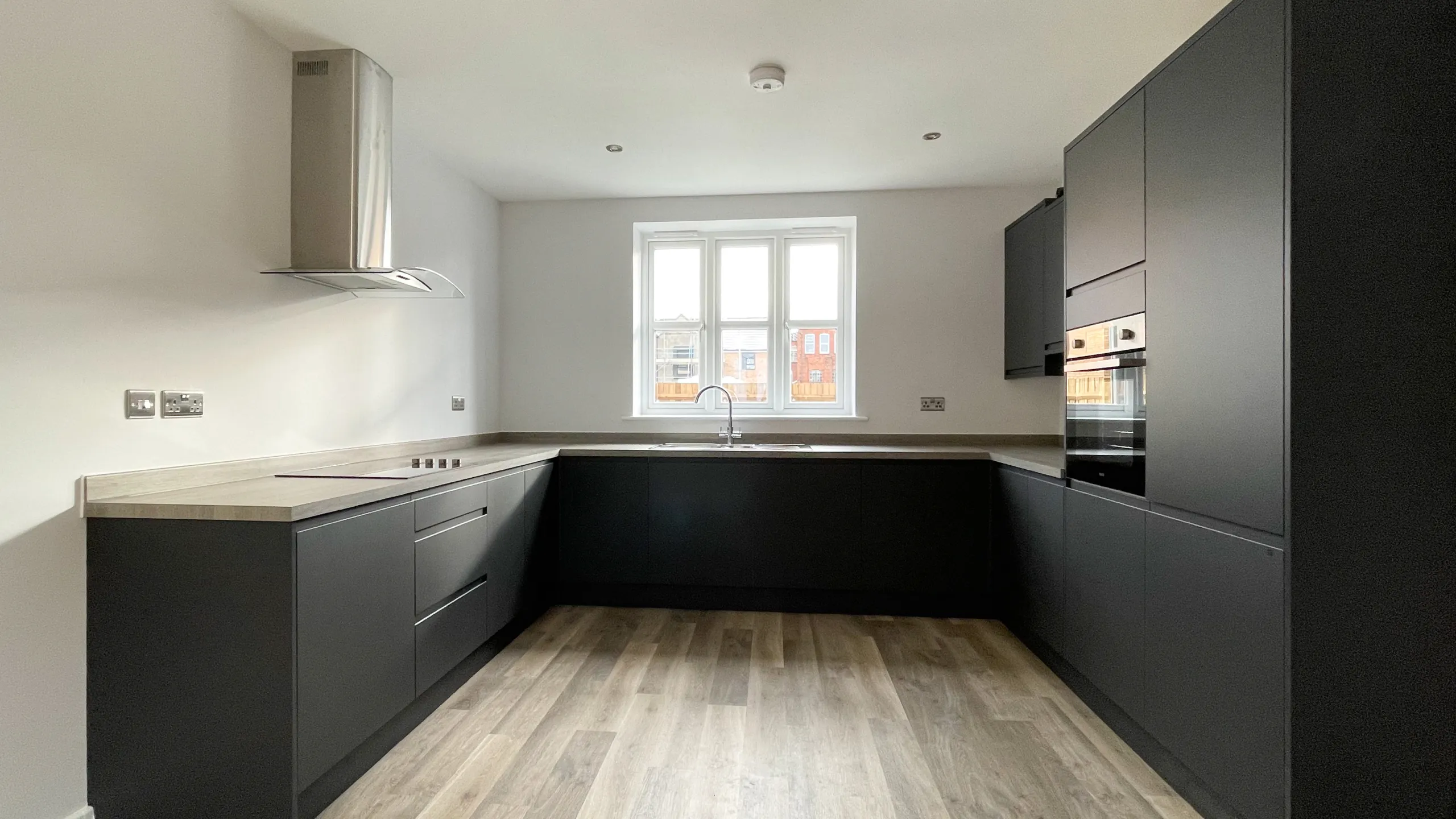 Modern U-shaped kitchen with black cabinets, light wood countertops, wooden floor, stainless steel oven, stovetop with hood, and a window above the sink.