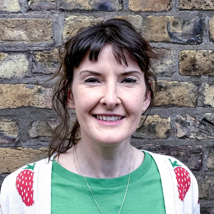 Smiling woman with dark hair wearing a green shirt and white sweater with strawberry patterns, standing in front of a brick wall.