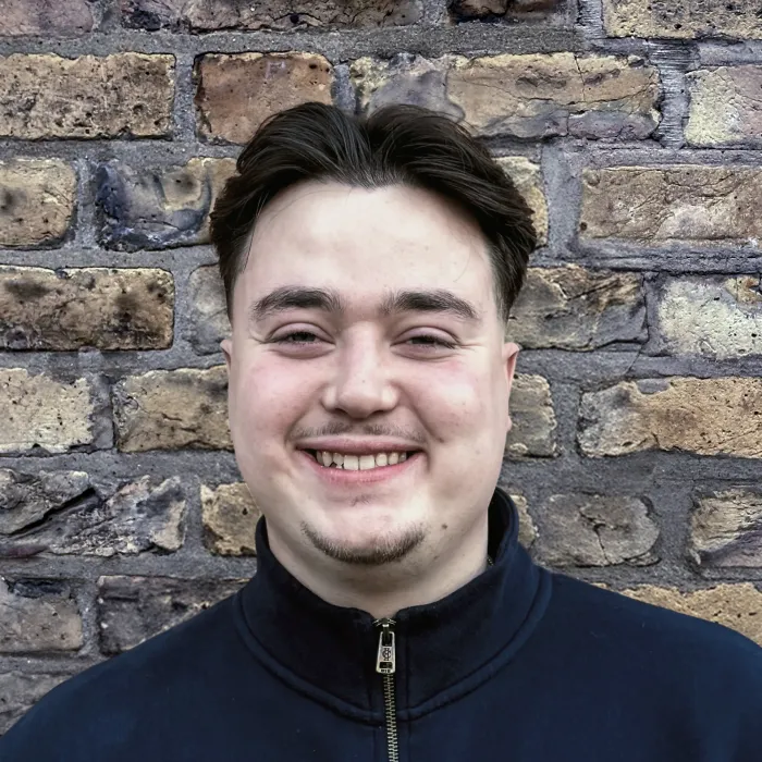 Young man with dark hair and light facial hair smiling in front of a brick wall.