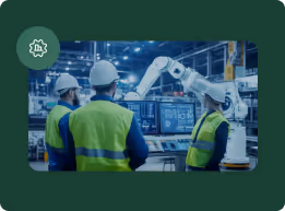 Three workers in safety gear monitoring robotic arm operations on computer screens in an industrial factory.