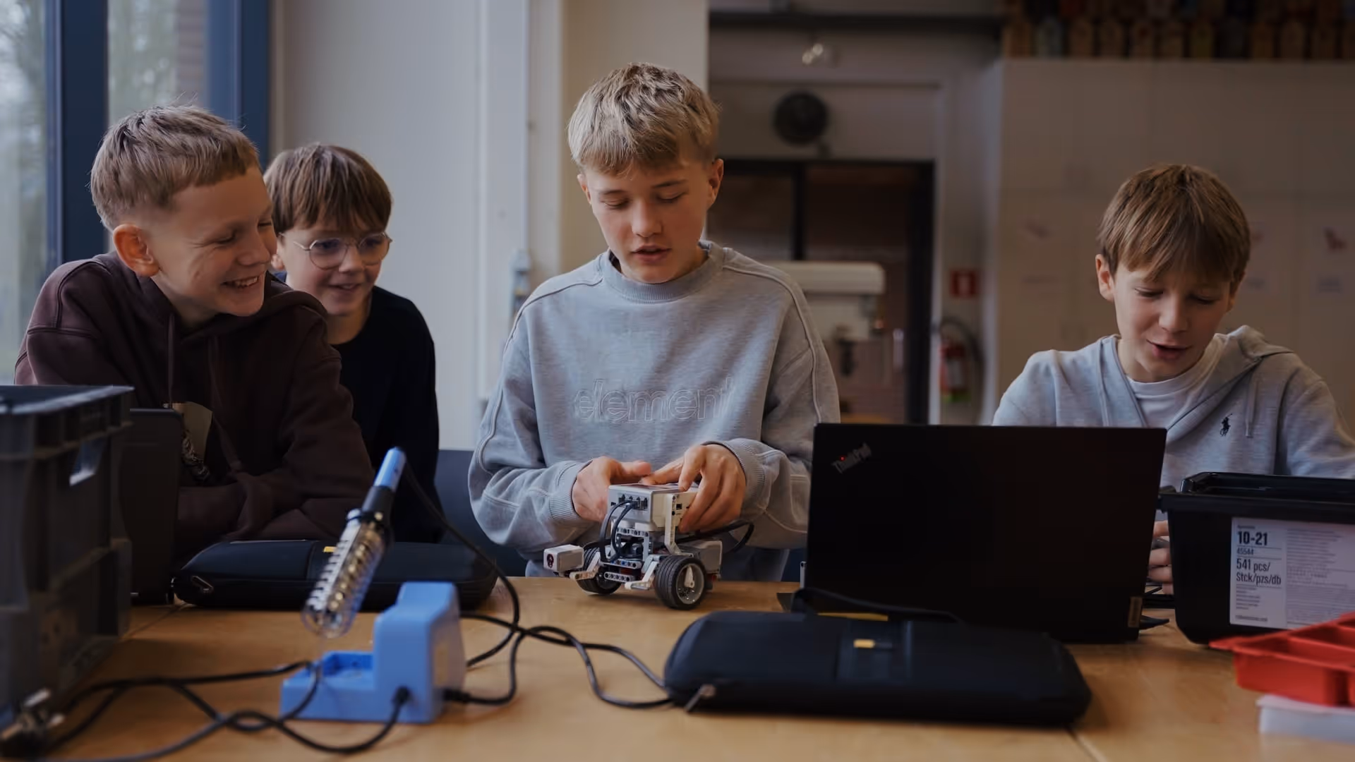 Een jongen in een grijs sweatshirt is intens bezig met het bouwen van een kleine robot aan een tafel met een laptop en soldeerapparatuur, en straalt nieuwsgierigheid en concentratie uit.