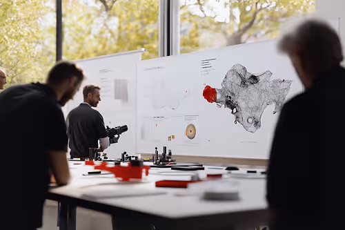Three people in a design studio working around a table with models and looking at architectural or urban planning diagrams on large boards by a window.