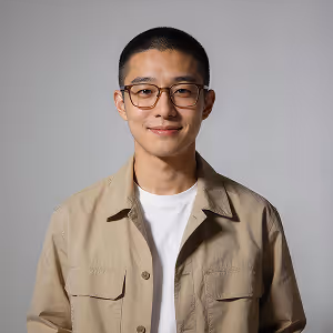 Young man with short hair and glasses wearing a beige shirt and white t-shirt against gray background.