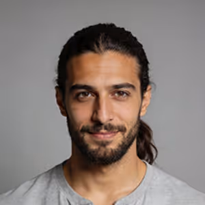 Portrait of a man with long dark hair tied back and a beard, wearing a gray shirt against a gray background.