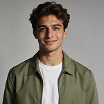 Smiling young man with curly hair wearing an olive green jacket over a white shirt.