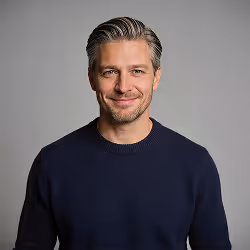 Smiling man with light brown hair in a navy blue sweater against a plain gray background.