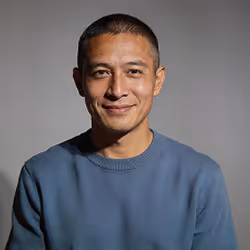Man with short hair smiling, wearing a blue sweatshirt, against a gray background.