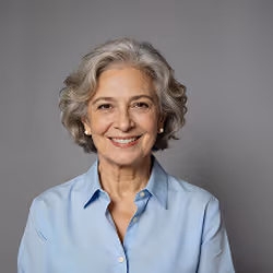Smiling older woman with gray curly hair wearing a light blue button-up shirt against a gray background.