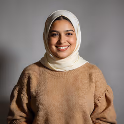 Smiling woman wearing a white hijab and a brown sweater against a gray background.